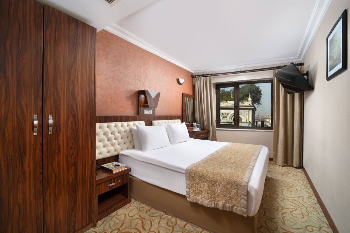 A hotel room with a large bed, wooden wardrobe, a nightstand, a bedside table, a window with curtains, and a wall-mounted TV.