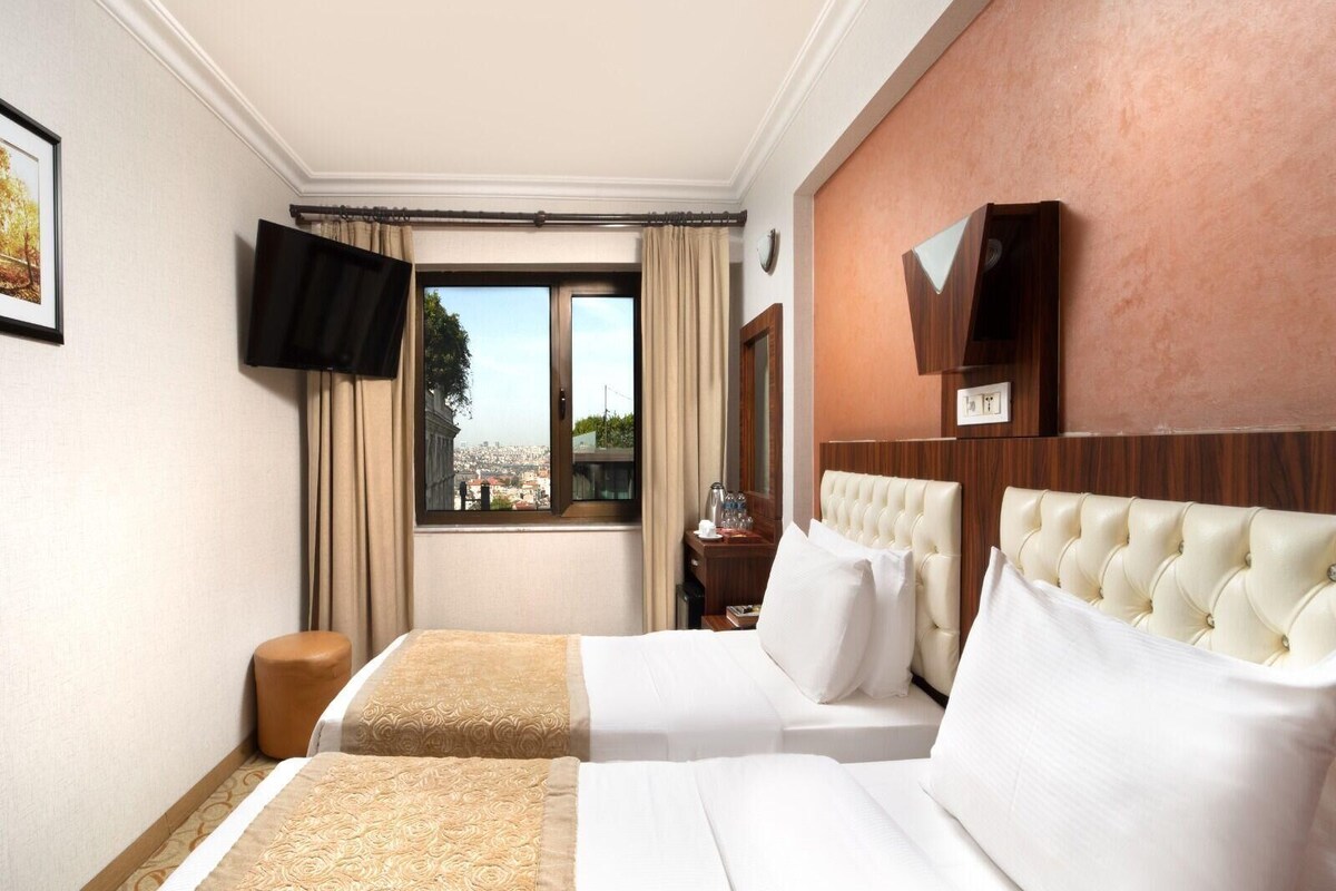 A hotel room with two beds, a television, a window with a view of the city, and a wooden headboard.