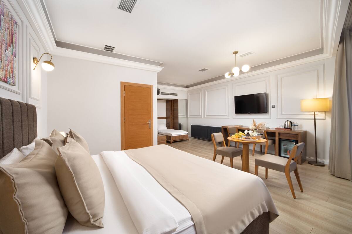 A hotel room with a large bed, a desk with a chair, a television, and a dining table with chairs.