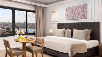 Une chambre d’hôtel avec un grand lit, une table à manger avec des fruits et du vin, une vue sur la ville et une œuvre d’art colorée accrochée au mur.