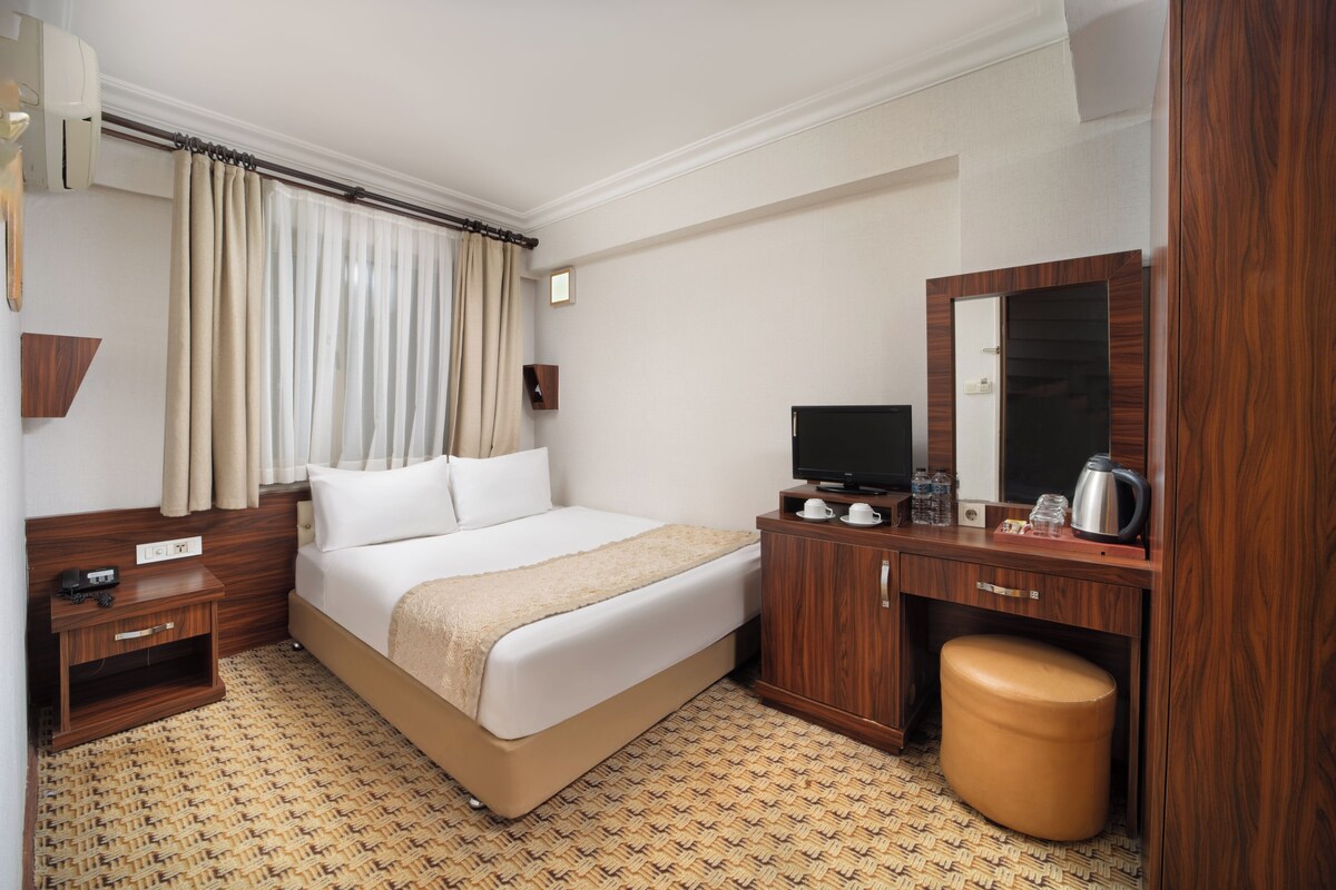 Una habitación de hotel con cama, mesita de noche de madera, televisión y un pequeño armario de madera con espejo.
