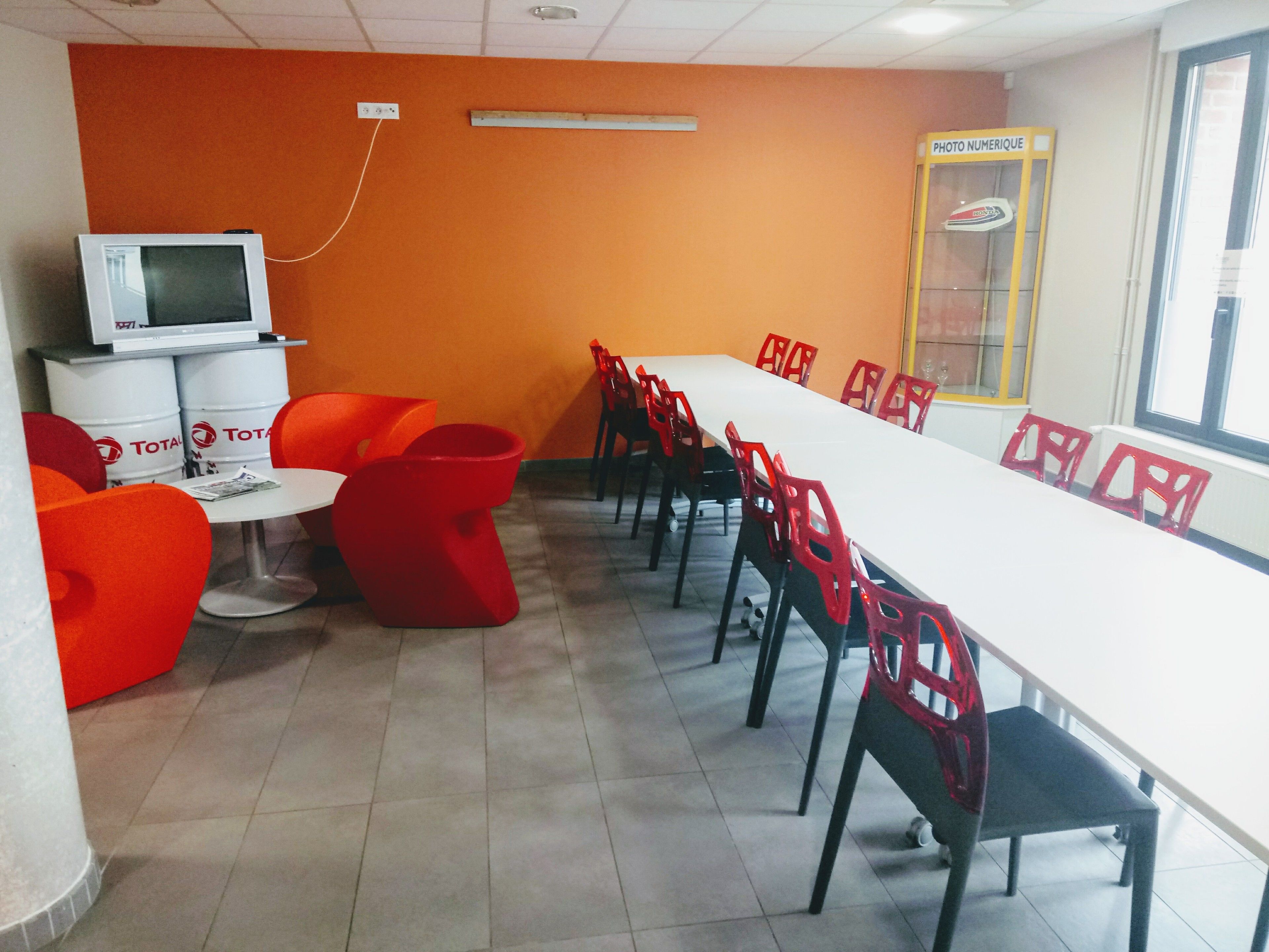 A conference room with a long white table, red and black chairs, a TV, and a 'PHOTO NUMERIQUE' sign.