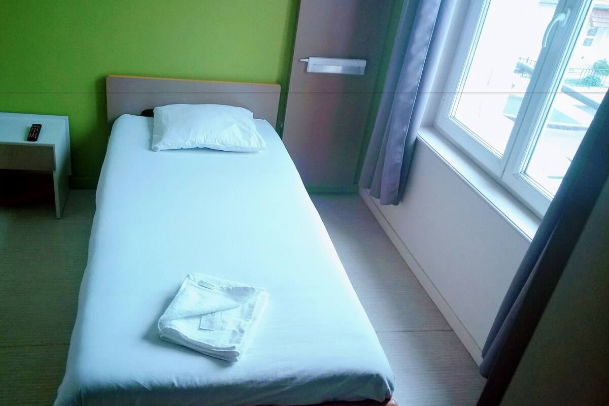 Ein Einzelbett mit weißer Bettwäsche, ein kleiner Nachttisch mit Fernbedienung und ein Fenster mit Vorhängen.
