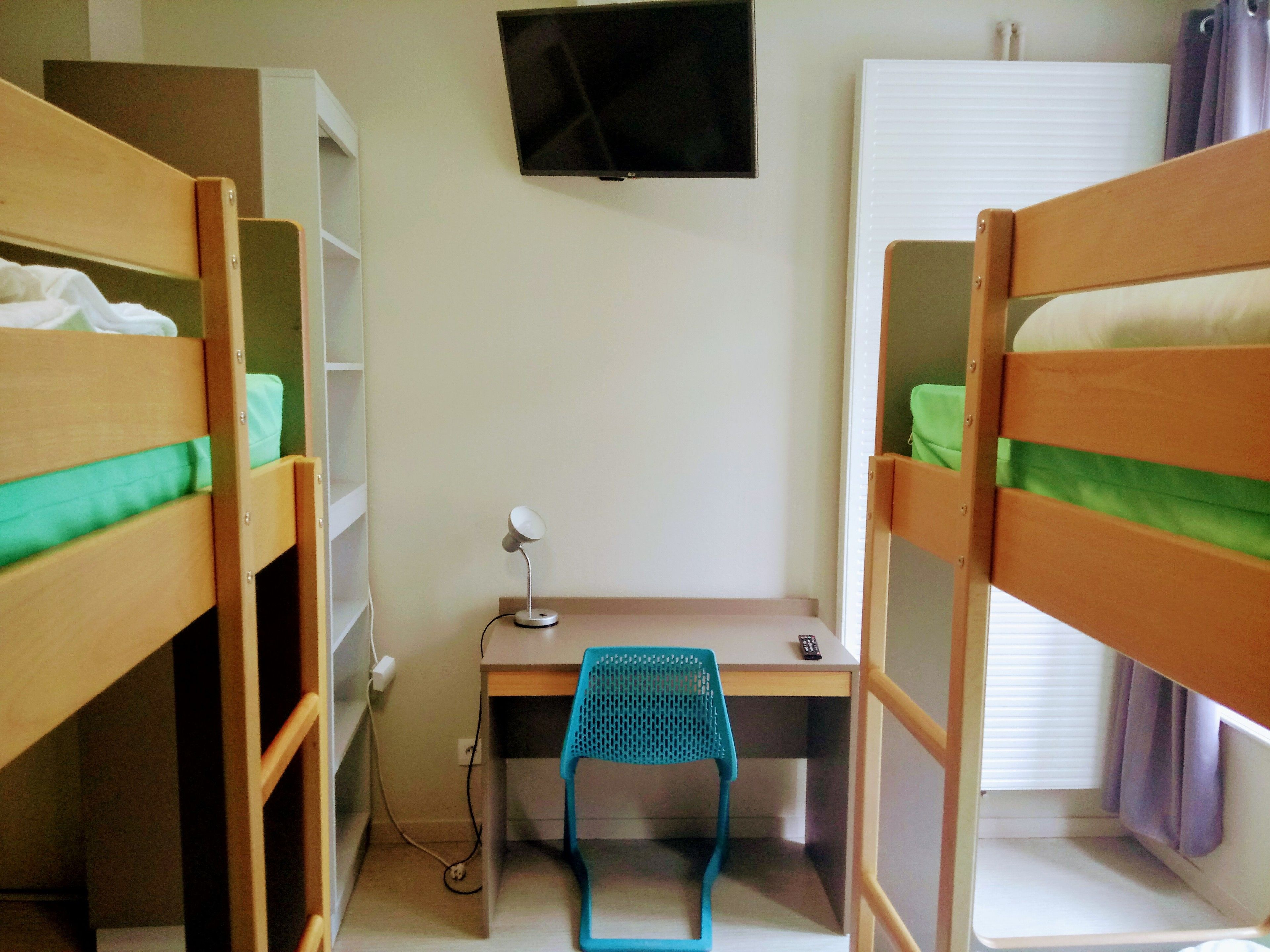 A dormitory room with bunk beds, a desk, a chair, a TV, and a window with curtains.
