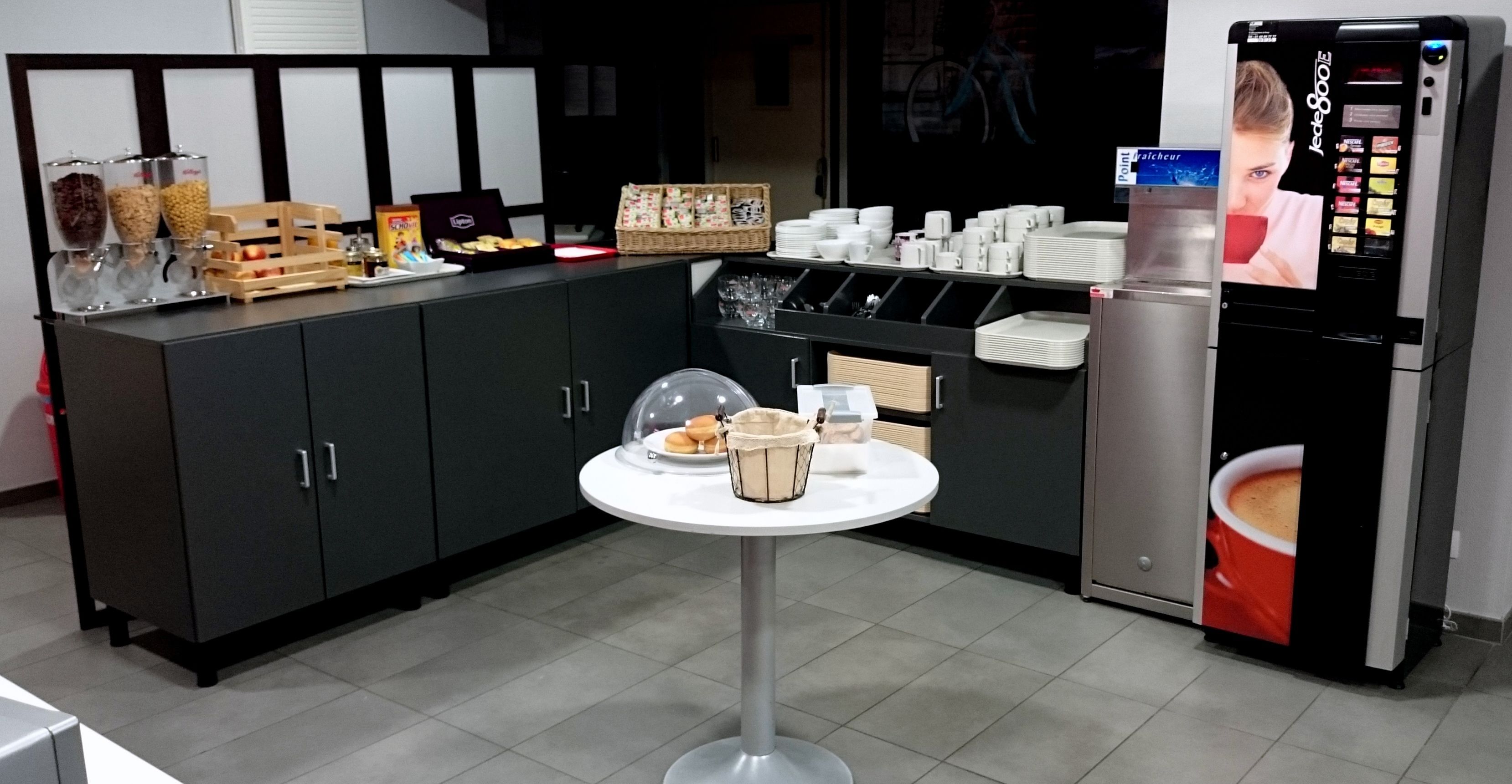 A kitchenette with a refrigerator, a coffee machine, and a small table with a basket and a plate.