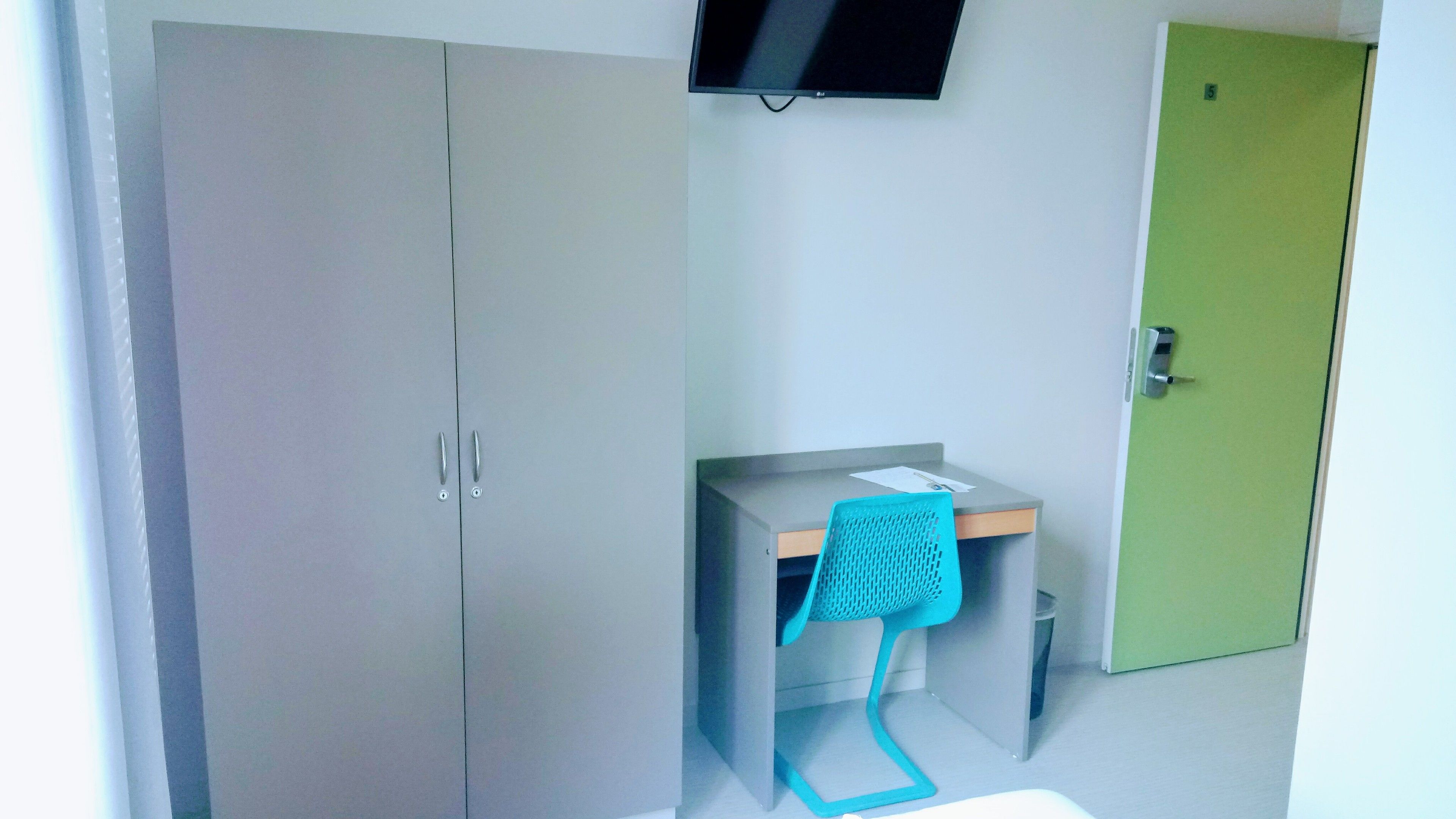 Een kamer met een grijze garderobe, een bureau met een blauwe stoel, een groene deur en een aan de muur bevestigde televisie.
