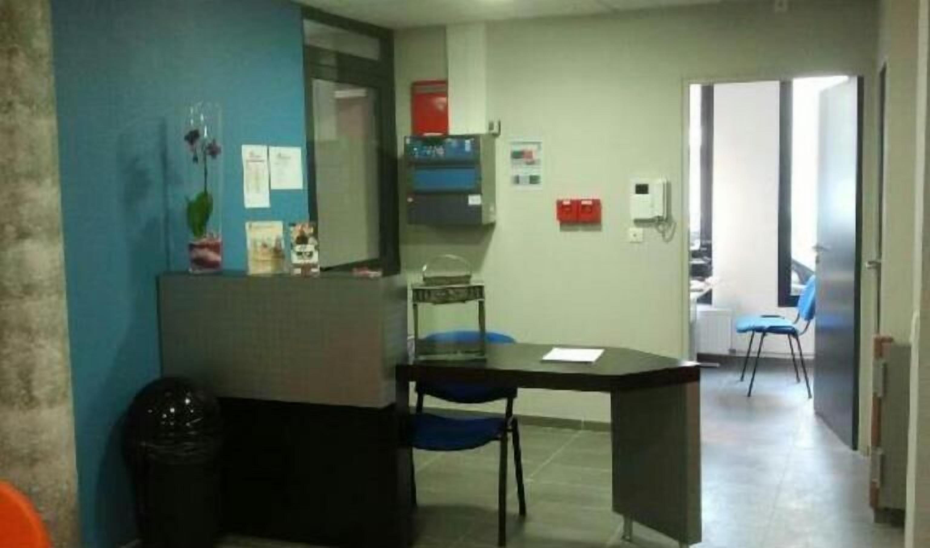 A small office with a desk, chair, and a trash bin. There is a blue wall with some decorations and a door leading to another room.