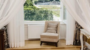 Een kamer met uitzicht op een grote tuin en een gebouw. Er staat een stoel met een kussen, een radiator en gordijnen.