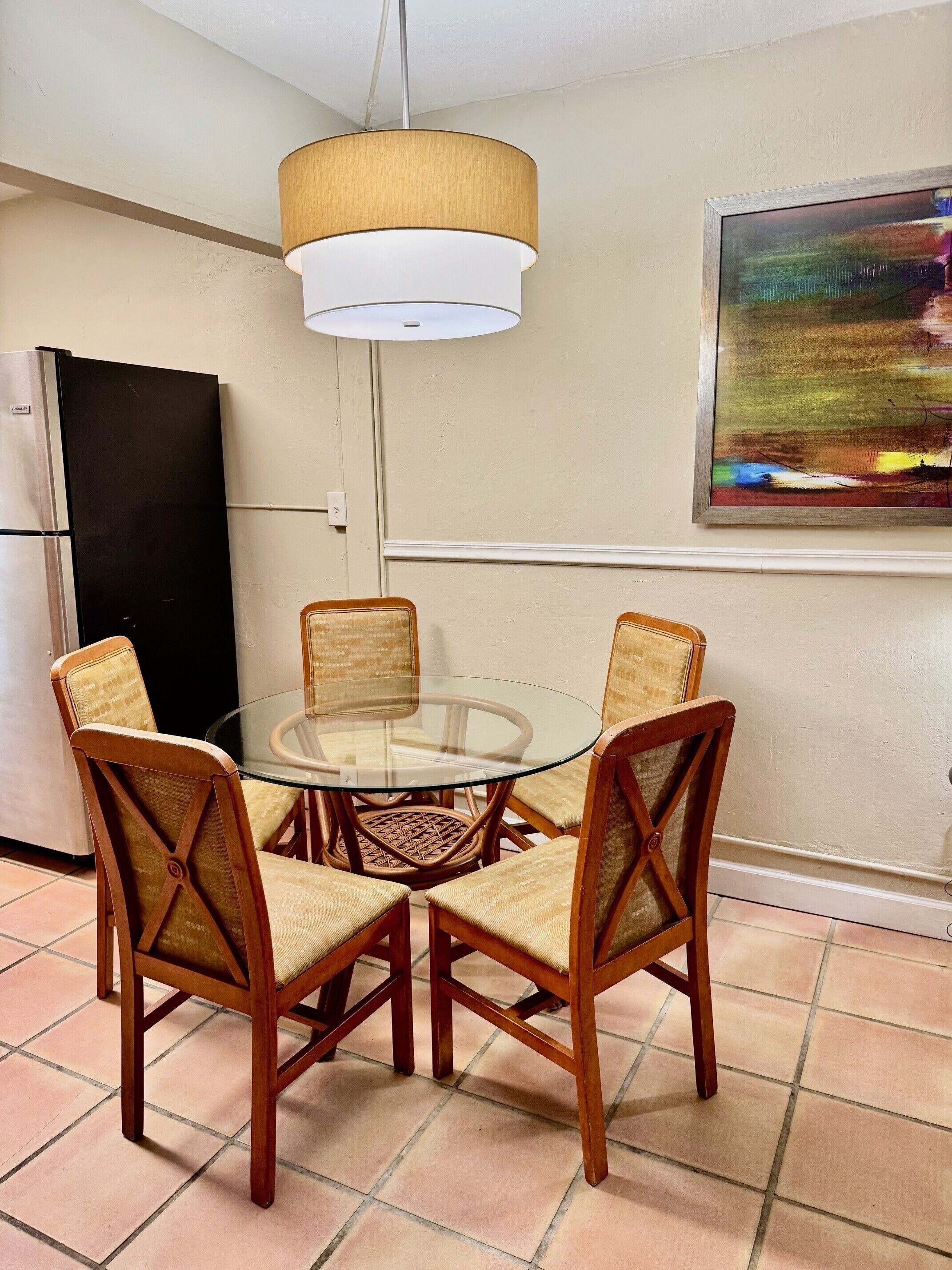 Un comedor con una mesa redonda de vidrio rodeada por cuatro sillas de madera con asientos de mimbre, un refrigerador negro y una pared color crema con un cuadro abstracto de colores.