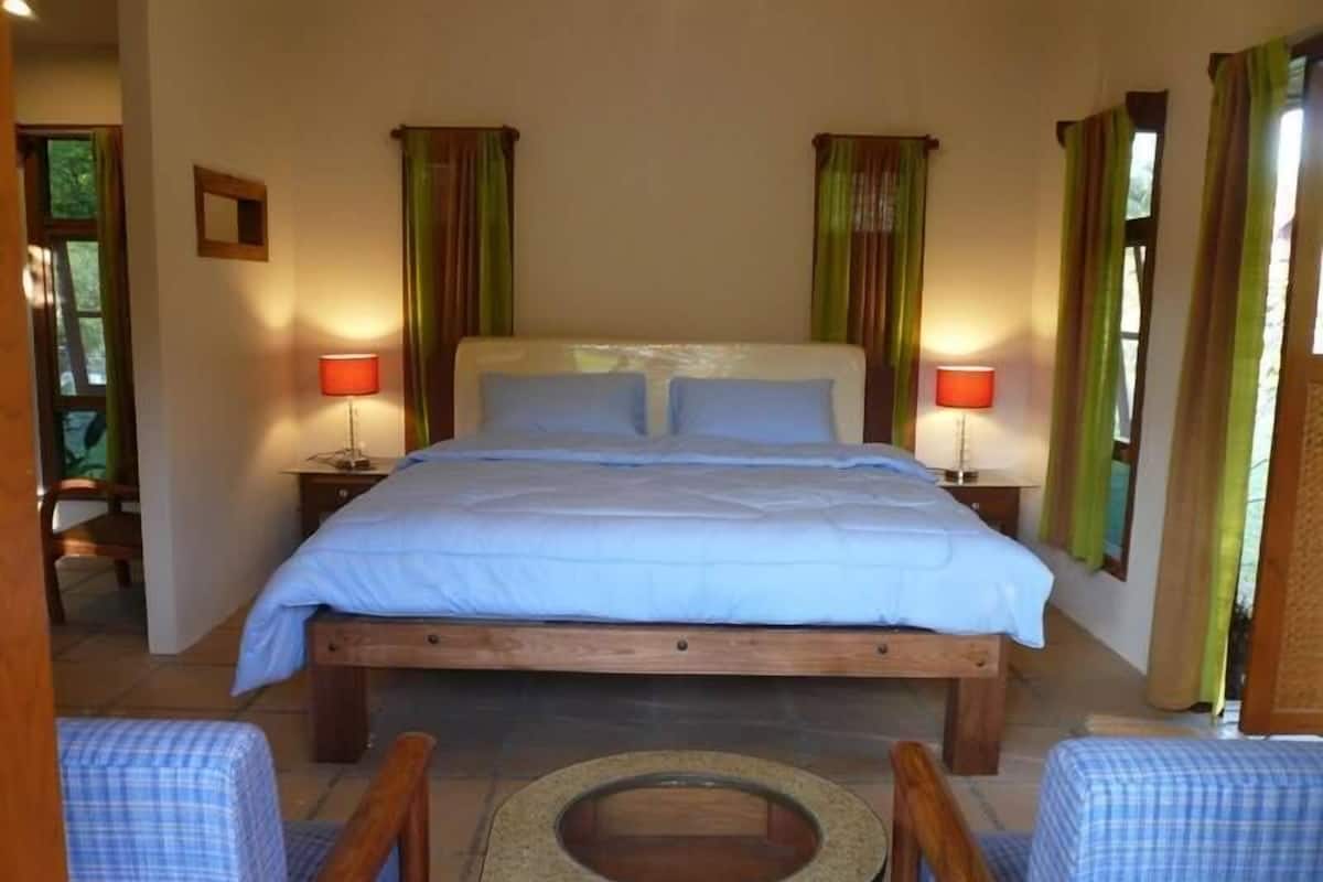A bedroom with a wooden bed, two bedside tables with lamps, a small window with curtains, and a view of the outdoors.