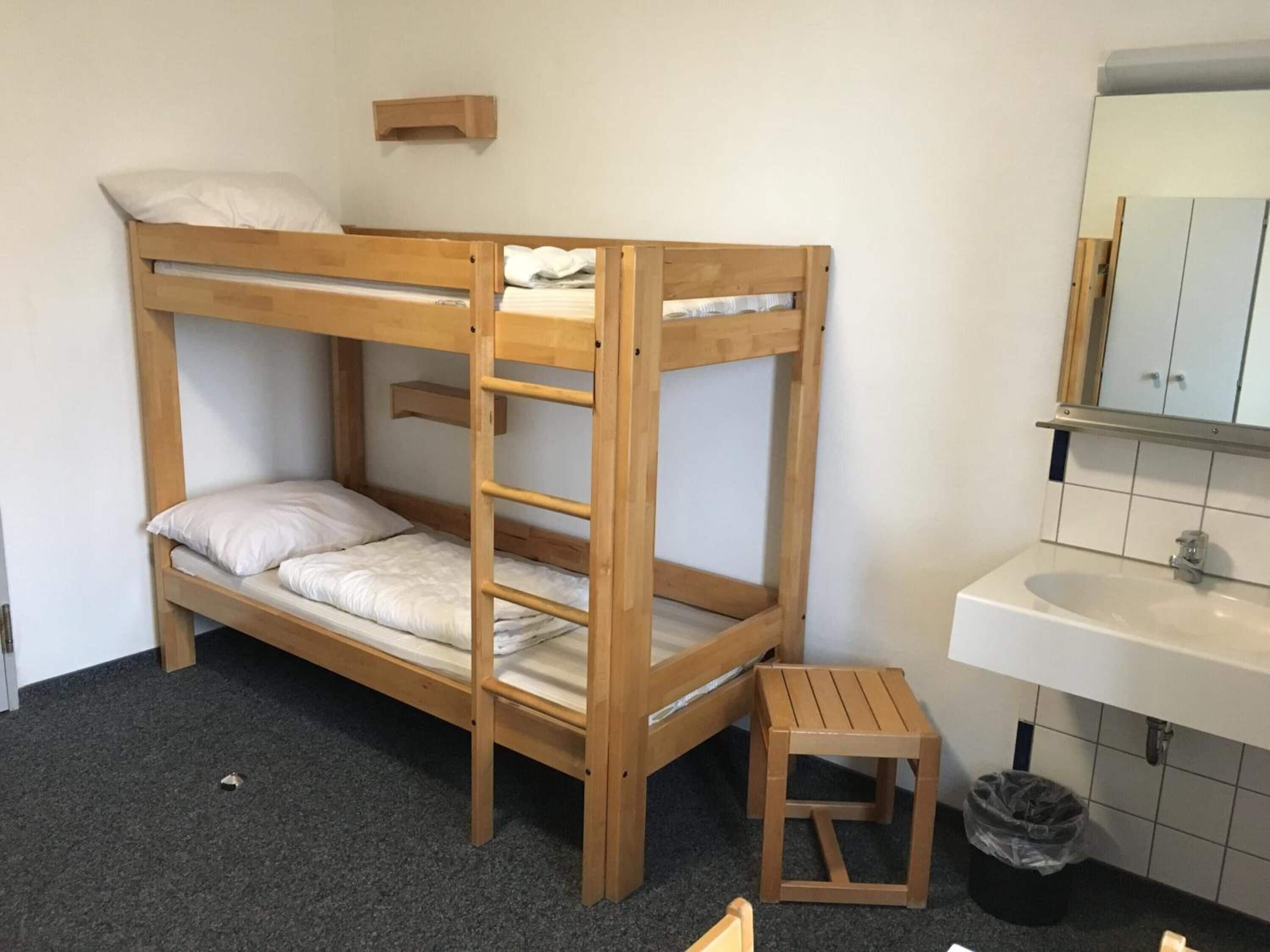 A bunk bed with a wooden ladder, a small wooden stool, a white sink, and a mirror above the sink.