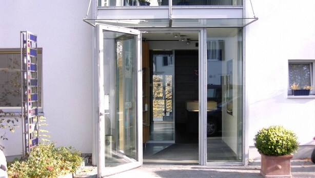 Un bâtiment moderne avec une entrée en verre, un petit jardin et une plante en pot à l’extérieur.