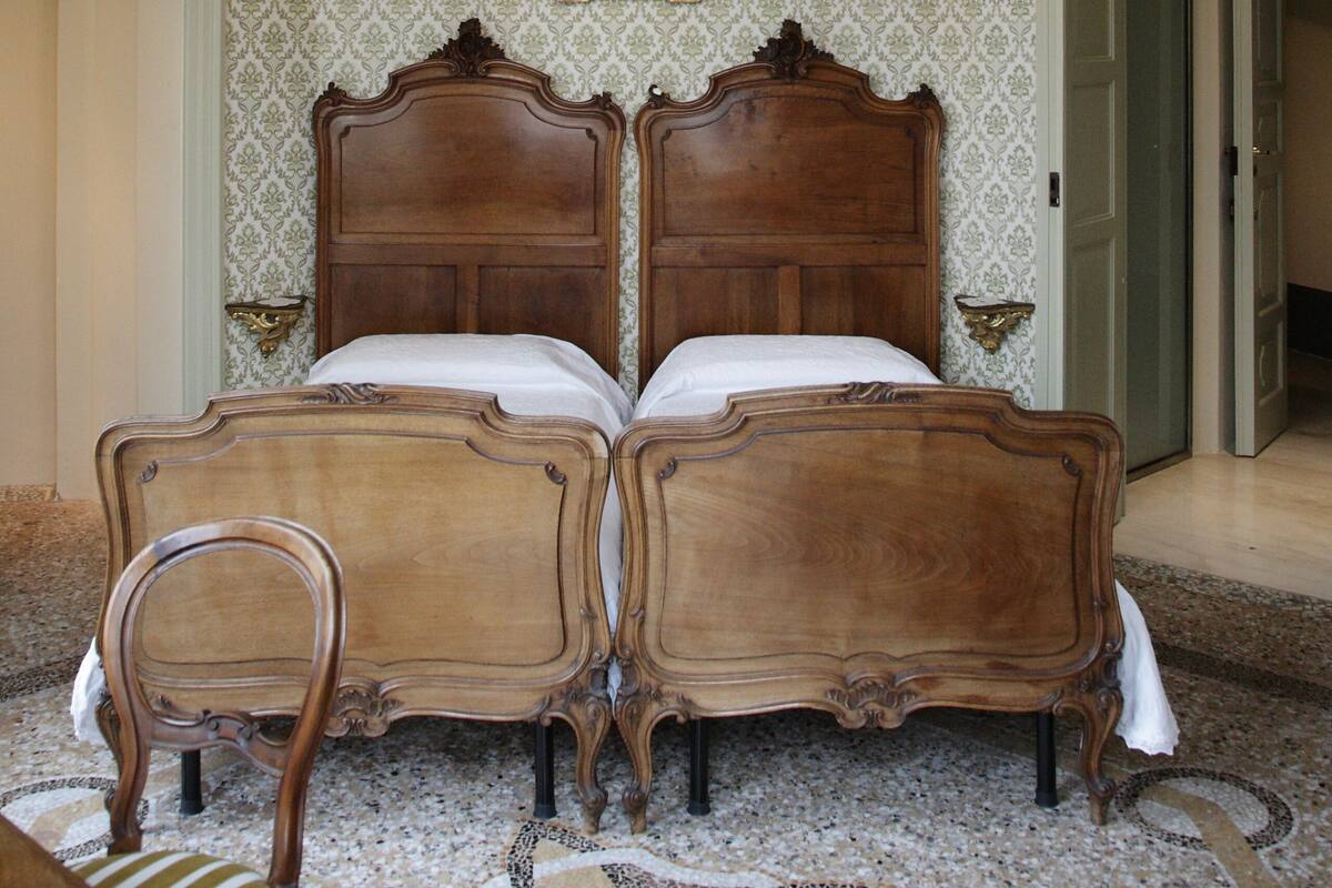 Ein Schlafzimmer mit zwei hölzernen Betten, einer Kristallleuchter, gemustertem Wandpapier und einem gerahmten Bild an der Wand.