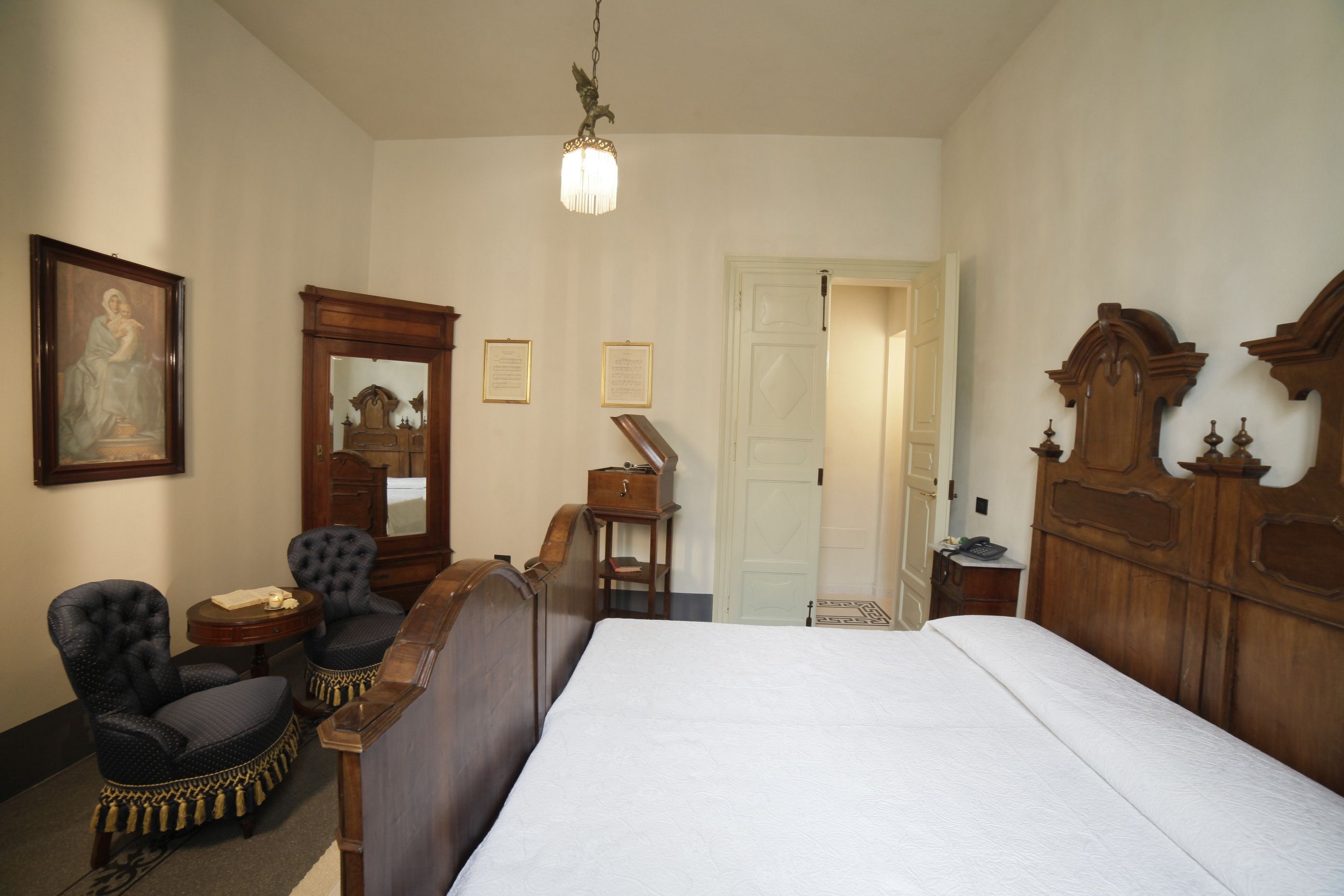 Un dormitorio con una cama grande, dos sillones tapizados, un aparador de madera, un espejo y un cuadro enmarcado en la pared.