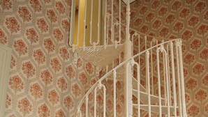 Un escalier en colimaçon avec une rampe en métal blanc, un luminaire jaune au sommet et un papier peint à motifs sur les murs.