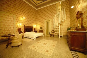 A spacious bedroom with a large bed, a sitting area with a chair, a wooden dresser, and a staircase leading to another level. - Residenza d'Epoca Regina d'Arborea (Oristano)