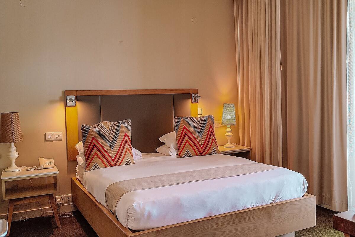 Ein Hotelzimmer mit einem großen Bett, zwei Nachttischen mit Lampen, einem Telefon und einem Fenster mit Vorhängen.