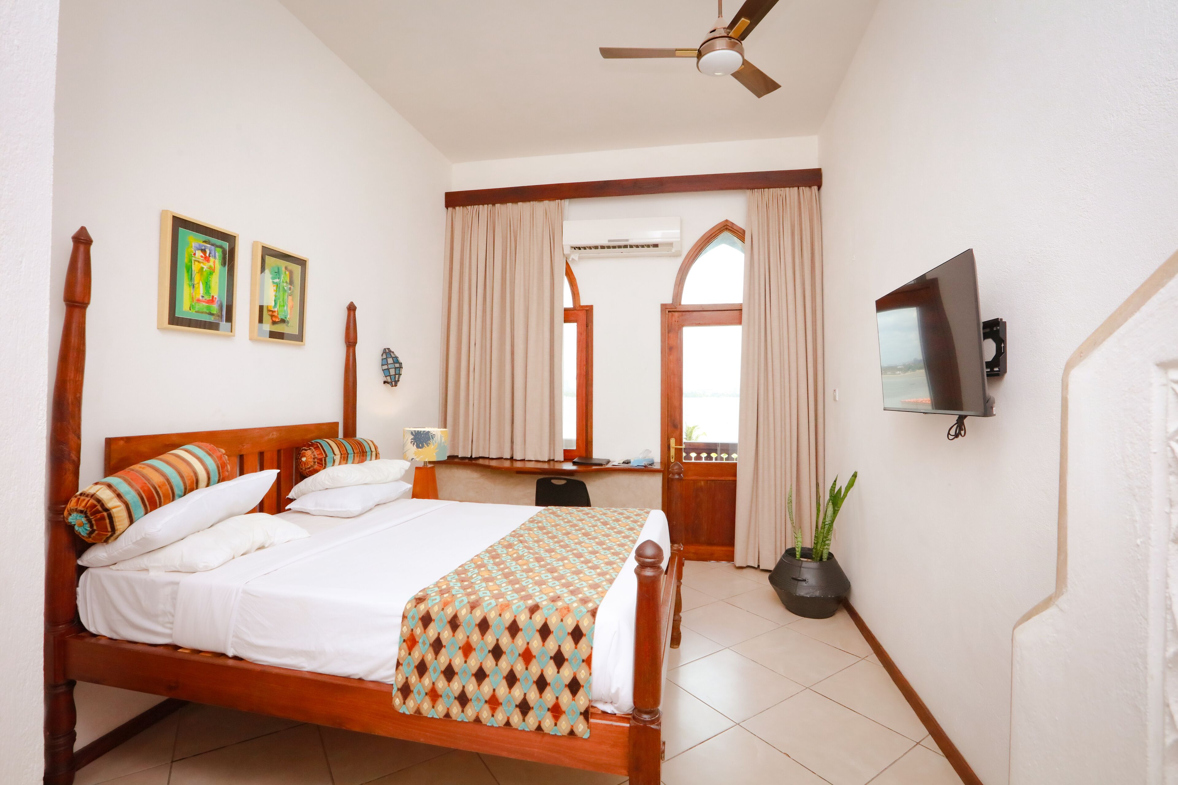 Una habitación de hotel con una cama de madera, un ventilador de techo, un televisor montado en la pared y una ventana con cortinas.