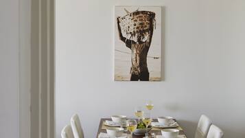 Comedor con una mesa de madera dispuesta para cuatro, sillas blancas y una obra de arte enmarcada sobre ella.