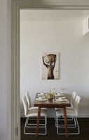 Comedor con una mesa de madera dispuesta para cuatro, sillas blancas y una obra de arte enmarcada sobre ella.