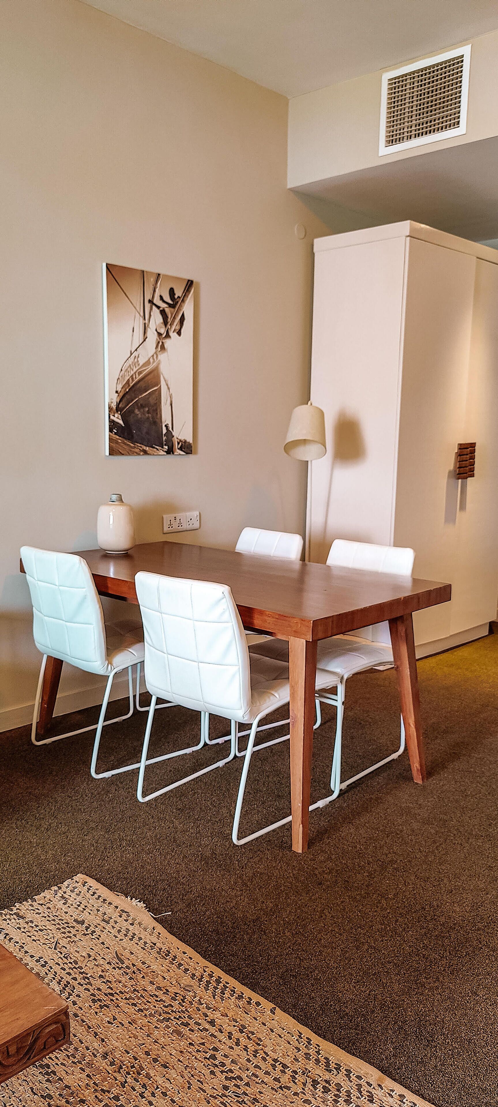Comedor con mesa de madera, sillas blancas acolchadas, lámpara de pared y un cuadro de un barco.