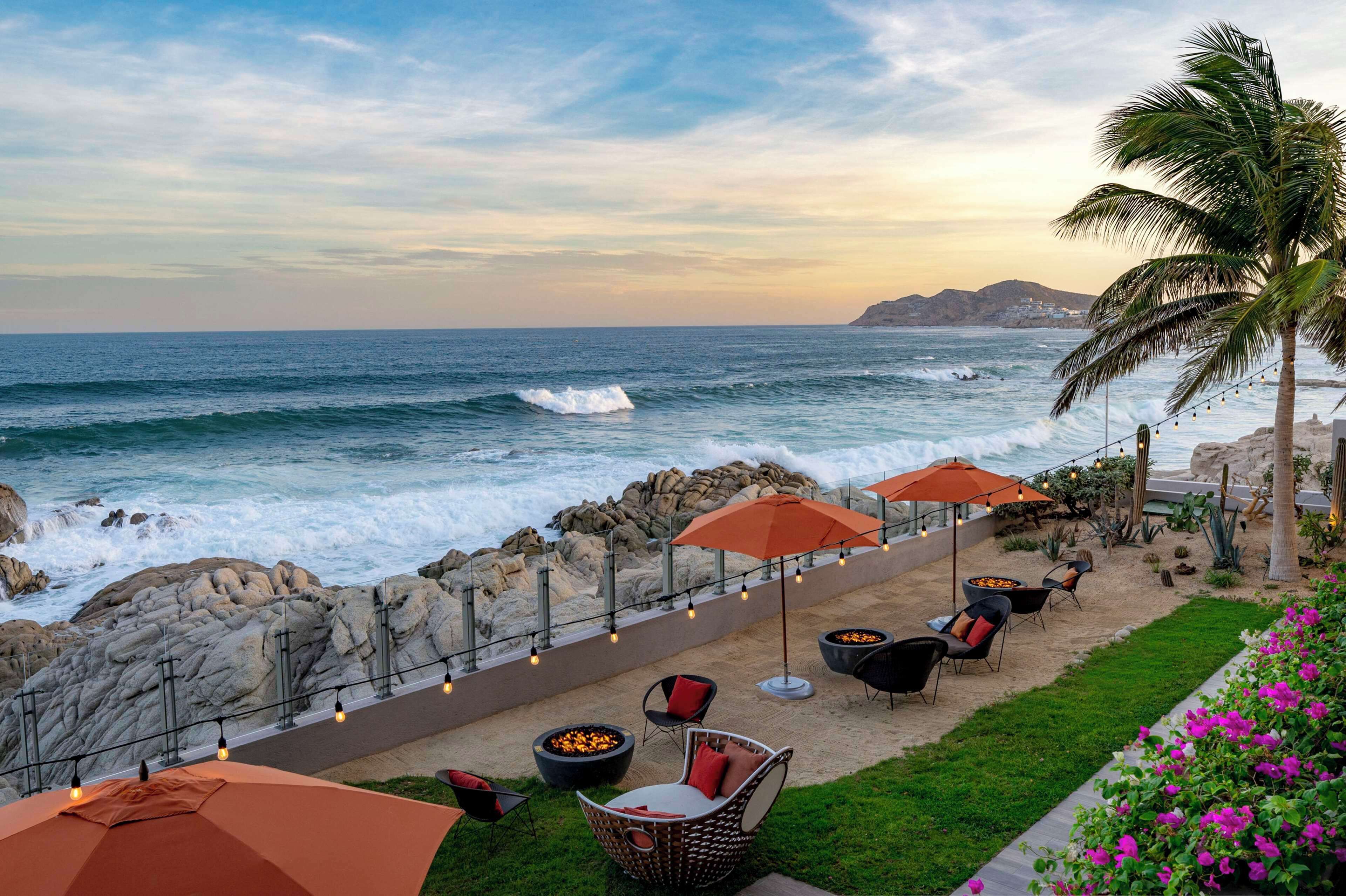 A coastal outdoor seating area with chairs, umbrellas, and a fire pit, overlooking the ocean.