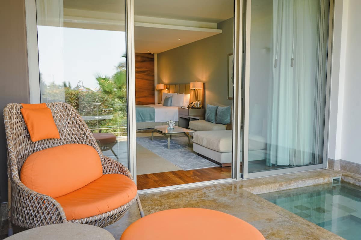 Una habitación de hotel moderna con un ventanal que da a un balcón, una silla de mimbre con cojín naranja, un sofá y una pequeña mesa.