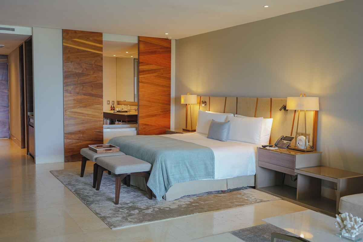 Une chambre d’hôtel moderne, dotée d’un grand lit, de tables de chevet, d’un bureau et d’une salle de bain visible à travers une porte ouverte.