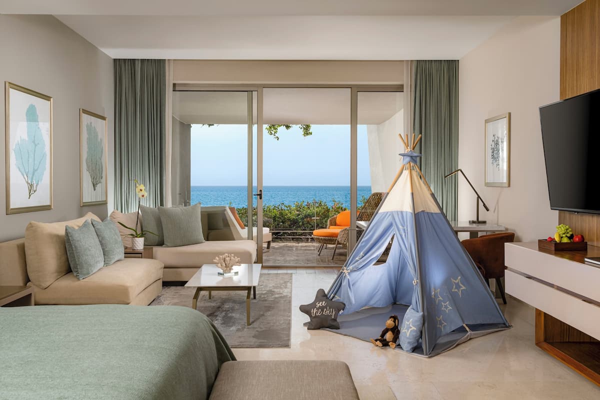 Una habitación de hotel moderna con una cama grande, un sofá, una mesa de centro, un televisor y vistas al mar a través de amplios ventanales.