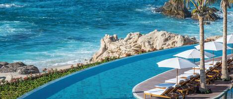 Una piscina infinita curvada con sillas de descanso y sombrillas, con vista a un litoral rocoso y al océano.