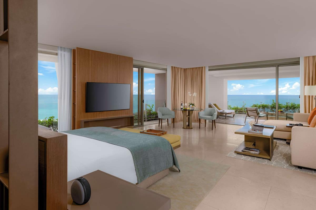 Una habitación de hotel moderna con una cama grande, un televisor, un balcón con vistas al océano y una zona de estar con una mesa y sillas.
