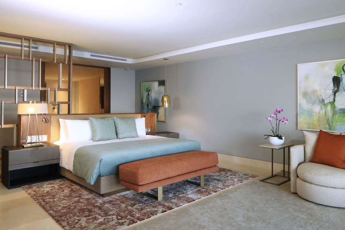 Una habitación de hotel moderna con una cama grande, una mesita de noche, un banco, una silla, una pequeña mesa con una planta y un cuadro en la pared.