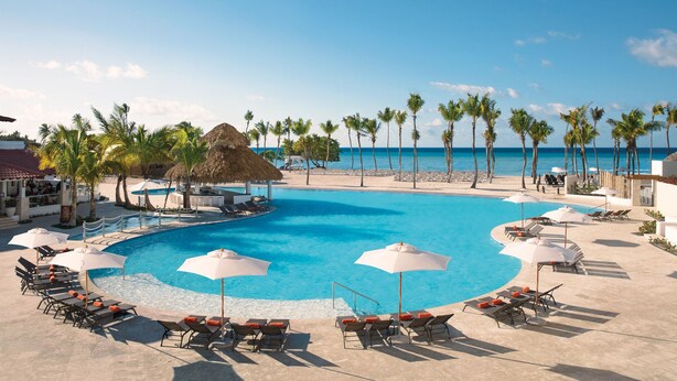 A pool area with lounge chairs and umbrellas, surrounded by palm trees and a view of the ocean. - Dreams Dominicus La Romana - All Inclusive (San Rafael del Yuma)