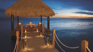 A wooden deck with a thatched-roof table and chairs, surrounded by lanterns, leading to a body of water. - Dreams Dominicus La Romana - All Inclusive (San Rafael del Yuma)