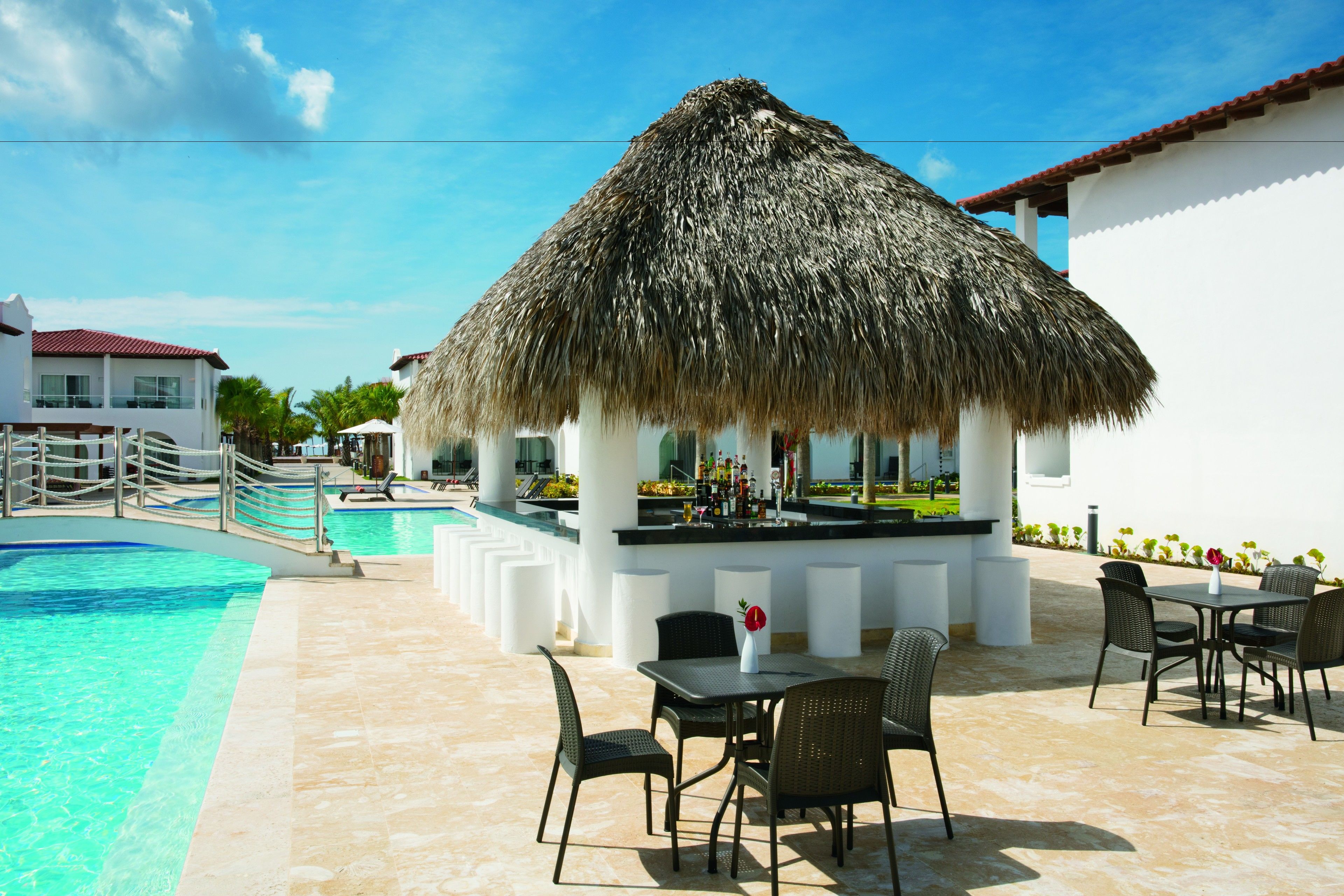 A poolside area with a thatched-roof bar, lounge chairs, and a dining table with chairs.