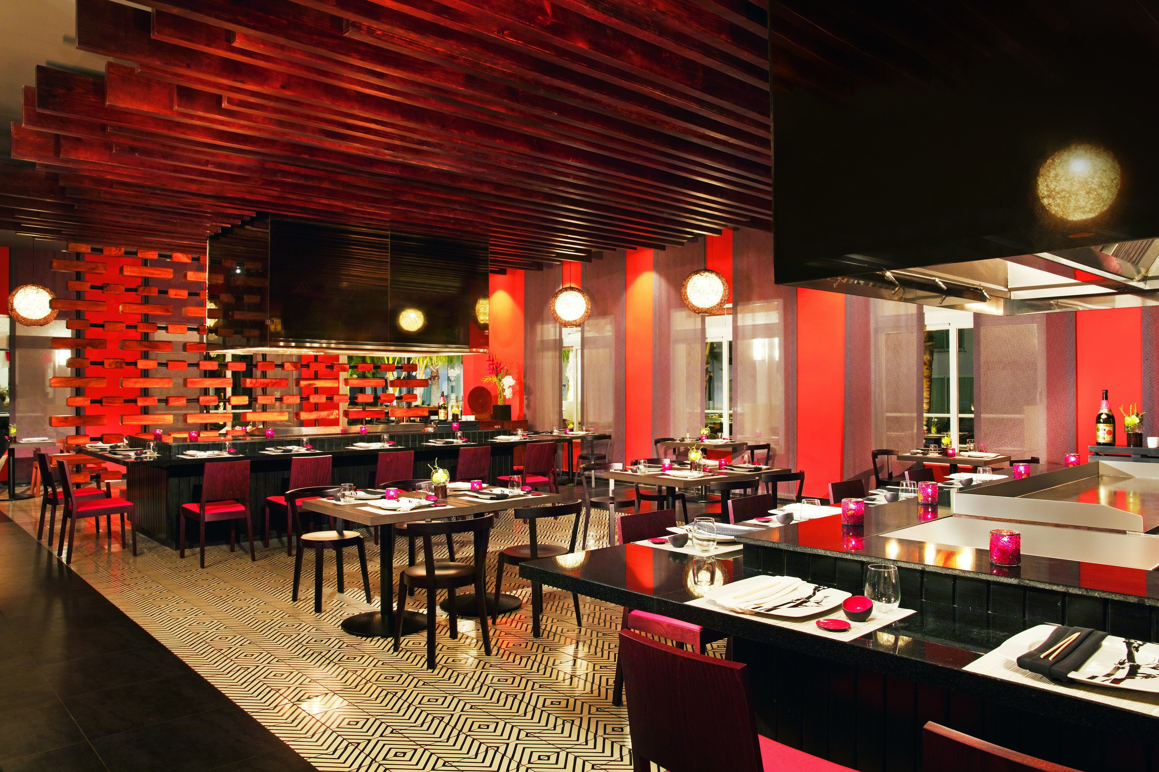 A modern restaurant interior with a red and black color scheme, featuring a central bar area, multiple dining tables, and a patterned floor.