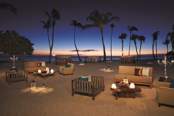 A beachside lounge with sofas, tables, and lanterns, overlooking the ocean and palm trees.