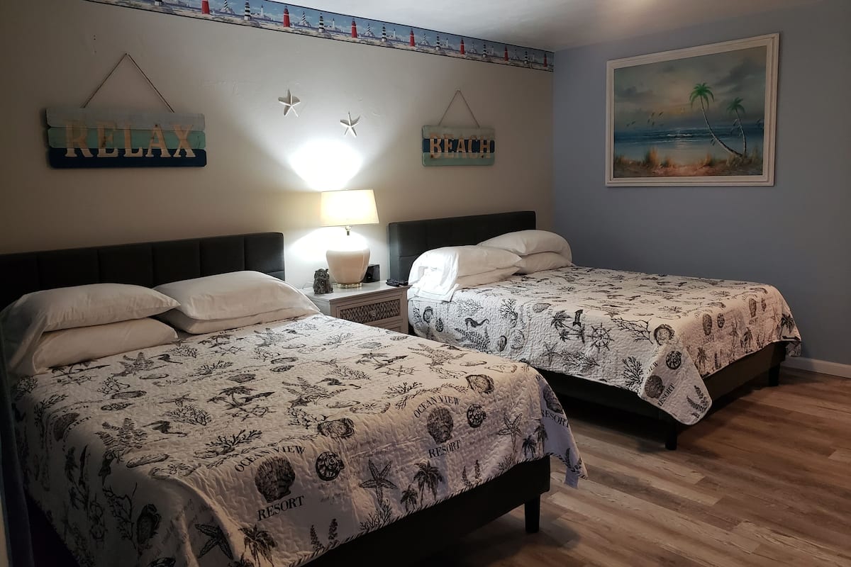 Una habitación con dos camas, una mesita de noche con lámpara, y adornos en la pared que incluyen un cartel que dice 'RELAX' y un cuadro con una escena de playa.