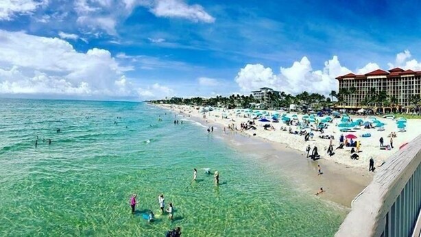 A beach with clear turquoise water, people swimming and sunbathing, and a hotel with red roofs in the background. - Ashley Brooke Beach Resort (Deerfield Beach)