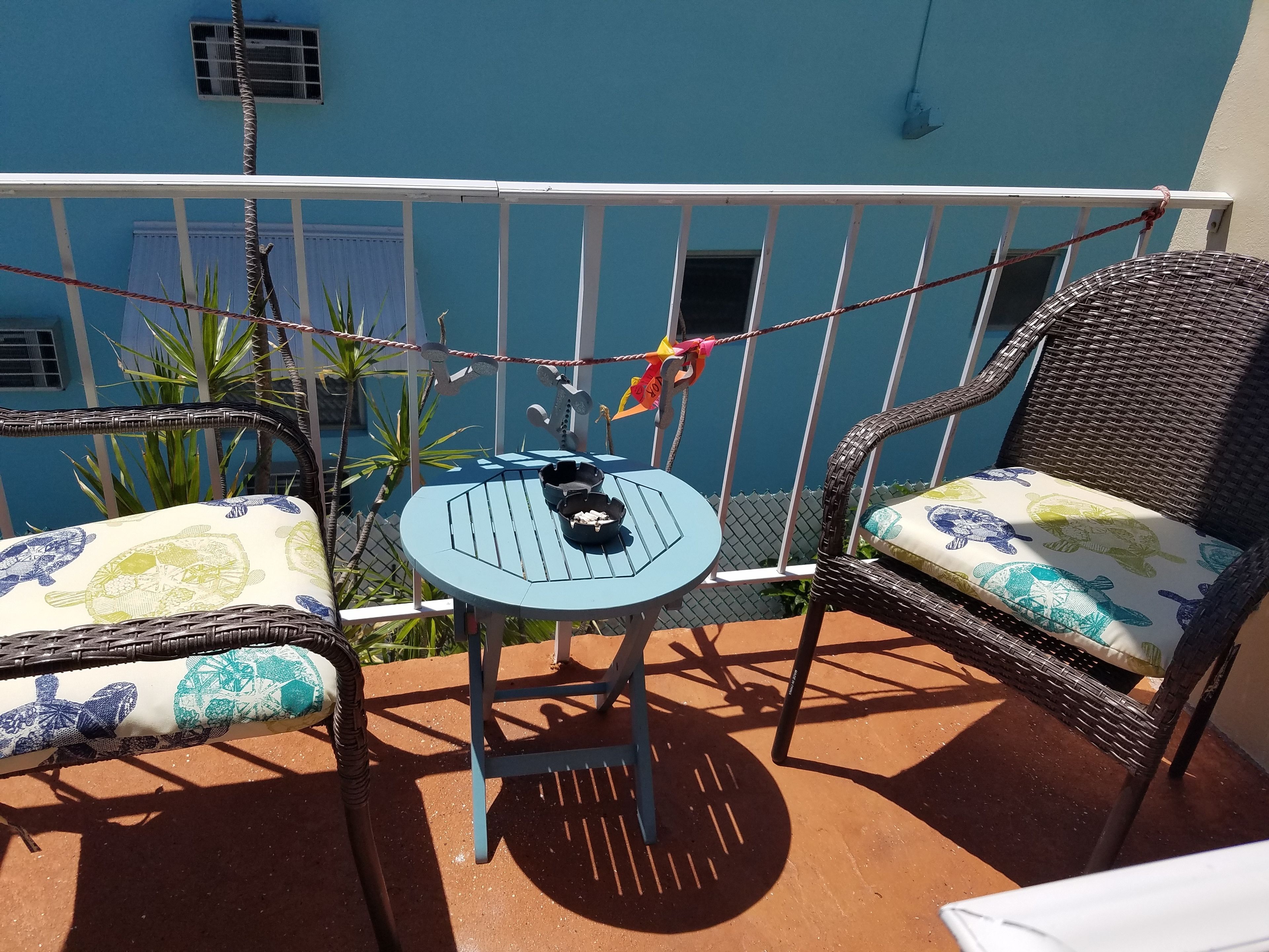 Ein kleiner Balkon mit Sitzgelegenheit im Freien, ausgestattet mit einem blauen Tisch und zwei Korbstühlen, einer davon mit einem Kissen mit Schildmuster.