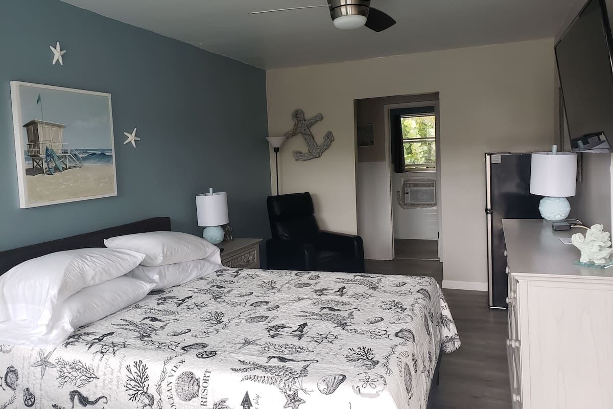 Un dormitorio con cama, escritorio, silla, ventilador de techo y un cuadro de una escena de playa.