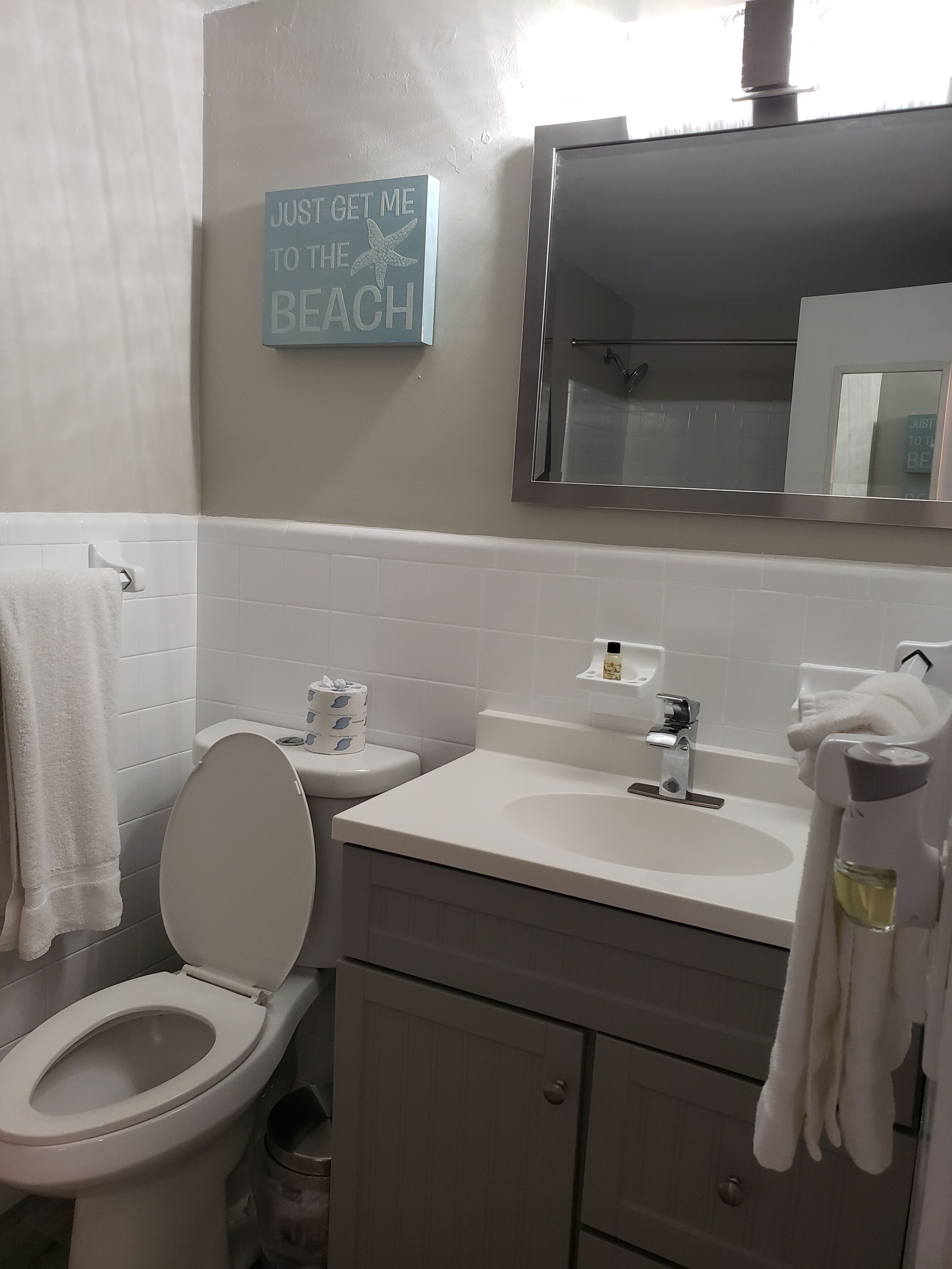 A bathroom with a toilet, sink, and mirror. There is a sign on the wall that says "Just get me to the beach."