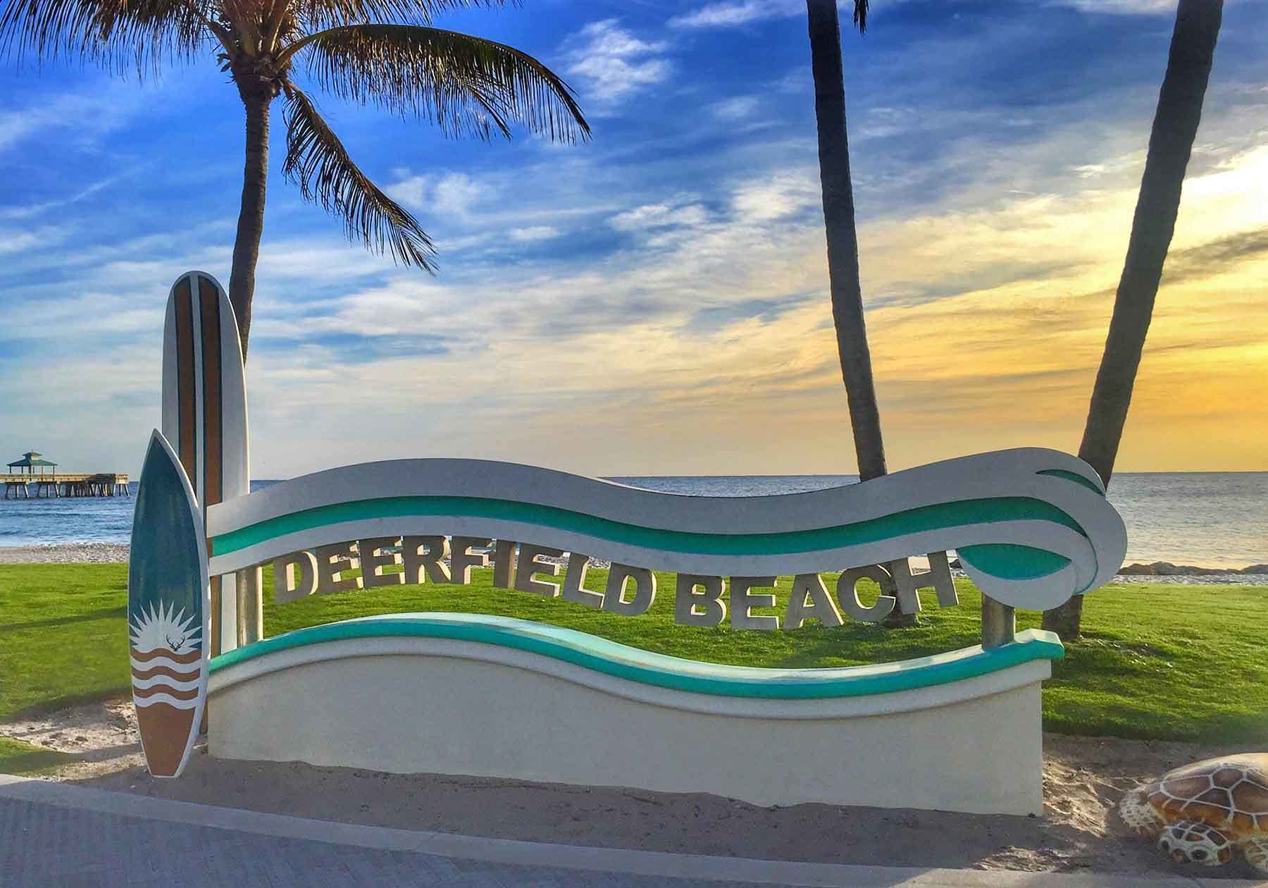 Un cartel de playa con el texto "Deerfield Beach" sobre un fondo de palmeras y una puesta de sol sobre el océano.