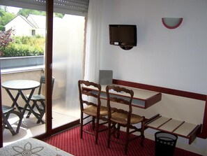 Une pièce avec une petite table et deux chaises, un lit, une fenêtre donnant sur des maisons et un téléviseur fixé au mur.
