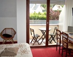 Une pièce comprenant un lit, un fauteuil en osier, une table à manger avec quatre chaises et une porte-fenêtre coulissante donnant sur un balcon extérieur d’où l’on peut admirer des maisons et la verdure.