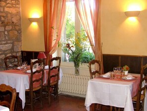 Un coin repas avec deux tables, chacune mise avec des assiettes, des verres et des couverts. Des chaises en bois sont disposées autour des tables, et une fenêtre avec des rideaux est visible.