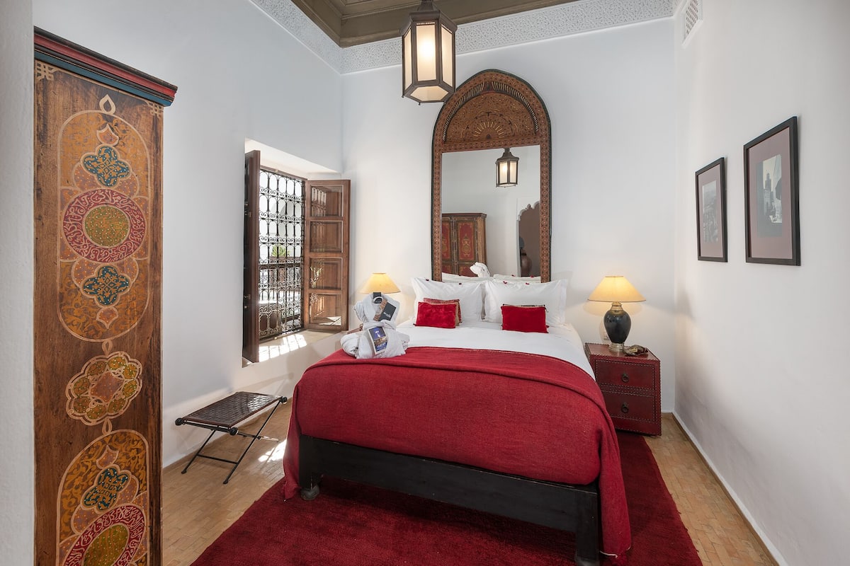 A bedroom with a large bed, a wooden wardrobe, a mirror, and a nightstand with a lamp.