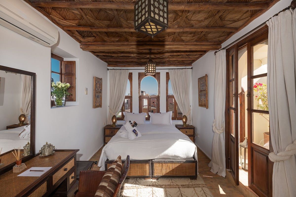 A bedroom with a wooden ceiling, a large bed, a bedside table, a mirror, and a window with curtains.