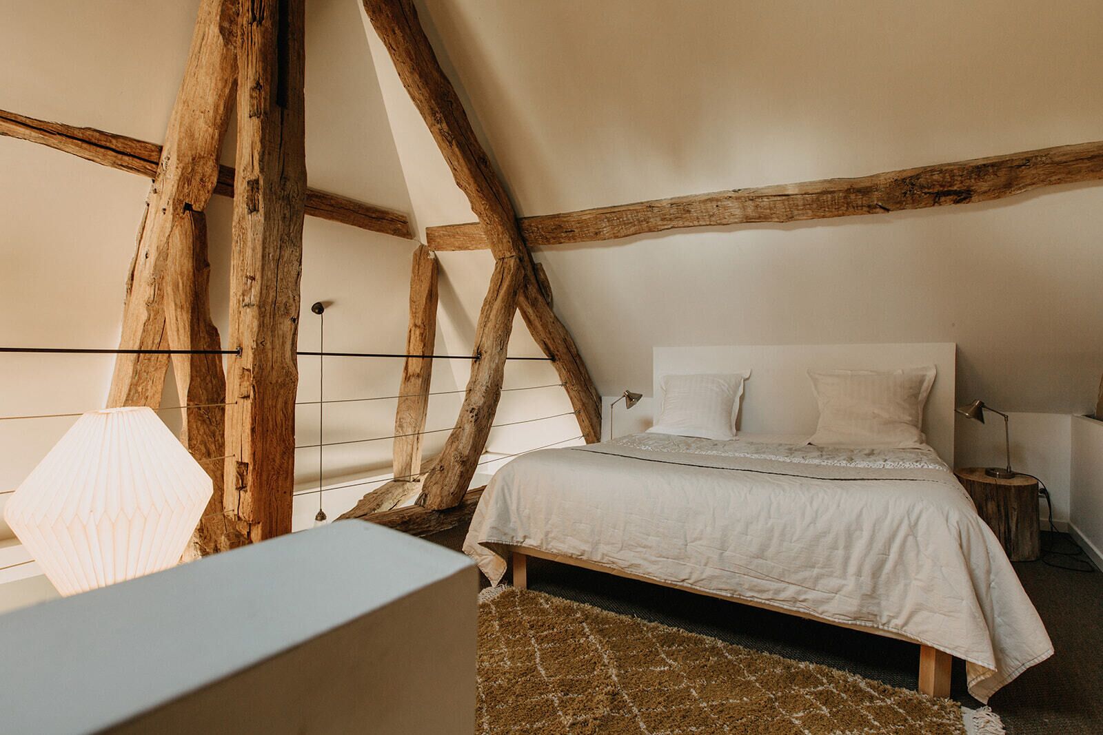 Un dormitorio con techo inclinado, vigas de madera a la vista, una cama con ropa de cama blanca, una mesita de noche y una lámpara.
