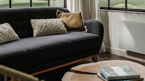 Een moderne woonkamer met een donkere bank, een ronde houten salontafel en uitzicht op een huis door grote ramen.